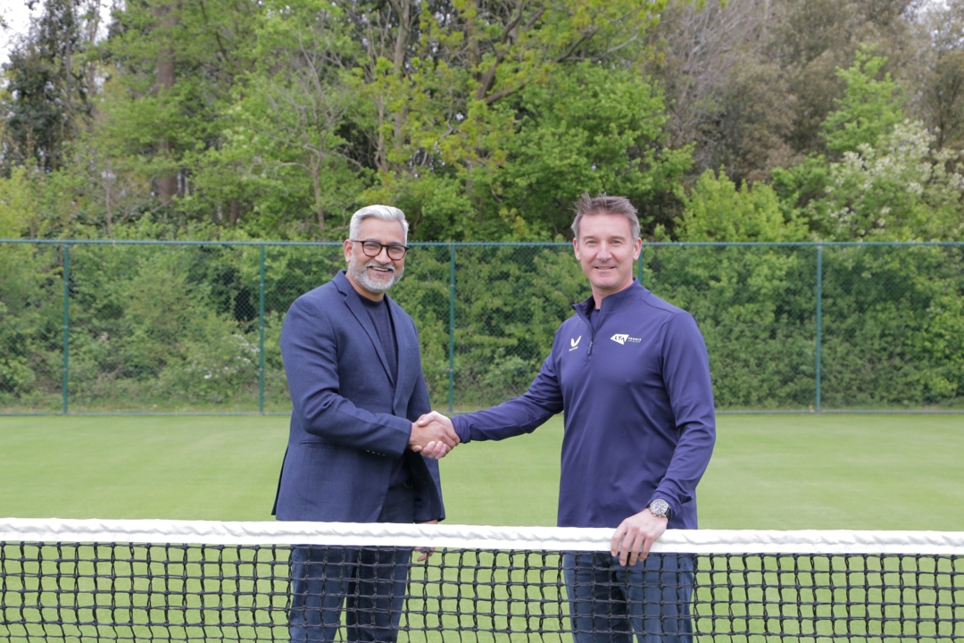 Two men standing behind a tennis net shaking hands and smiling. They are stood on grass, with green foliage in the background behind them
