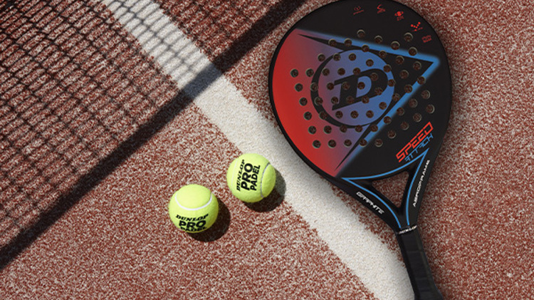 Two padel balls and a Dunlop padel bat on a clay-coloured padel court