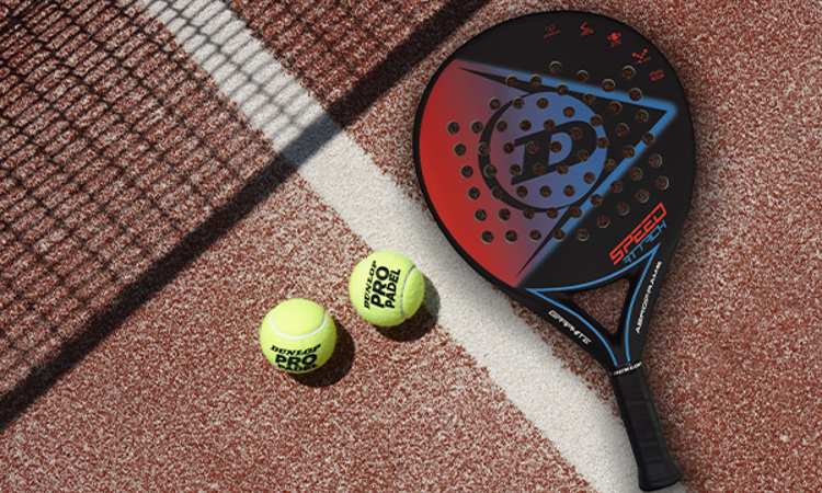 Two padel balls and a Dunlop padel bat on a clay-coloured padel court