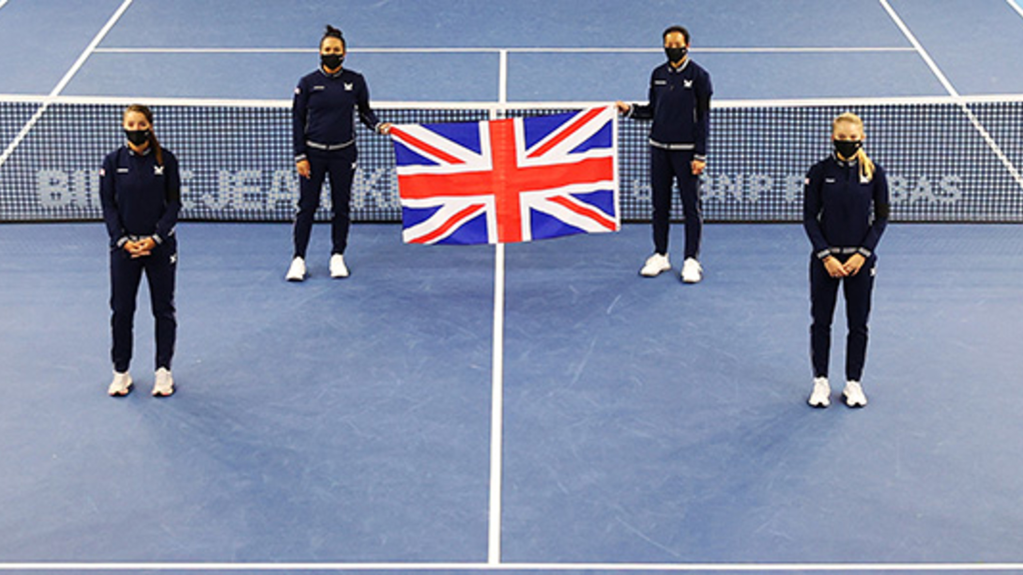 Six people standing a small distance apart on a tennis court with the back two holding a United Kingdom flag up