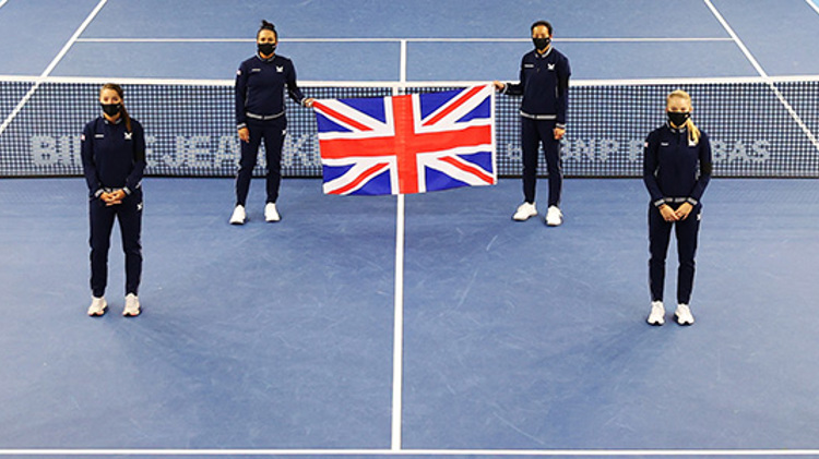 Six people standing a small distance apart on a tennis court with the back two holding a United Kingdom flag up