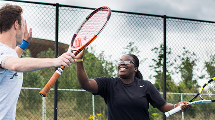 Two people high fiving while playing tennis with courtside fence in background