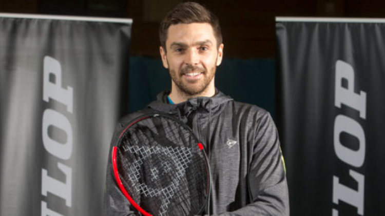 colin fleming  smiling for a picture  wearing all back with a racket in hand and dunlop  banners at the back