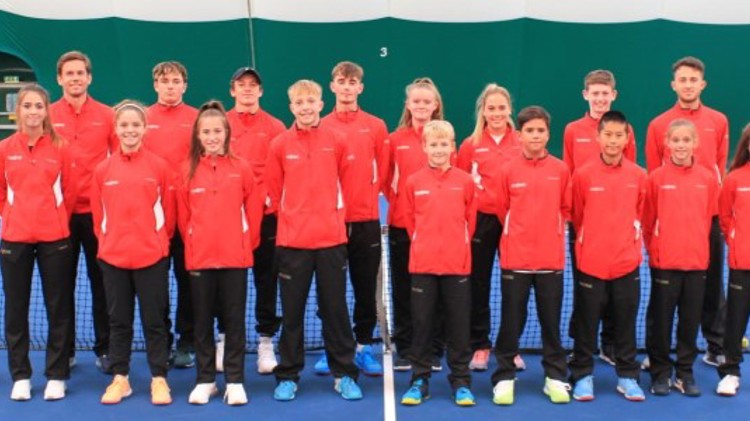Group picture of the junior team wales standing on court in front of the net wearing red jackets and smiling