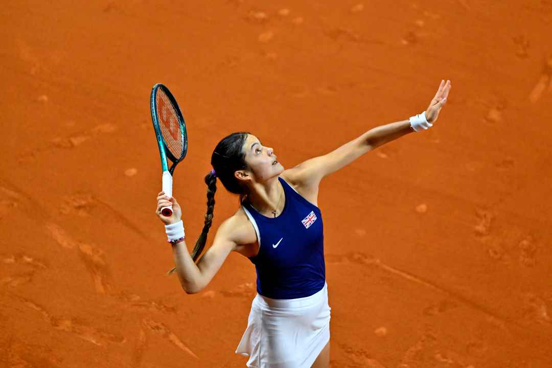 Emma Raducanu tossing a tennis ball in the air while preparing to serve on an orange clay court 