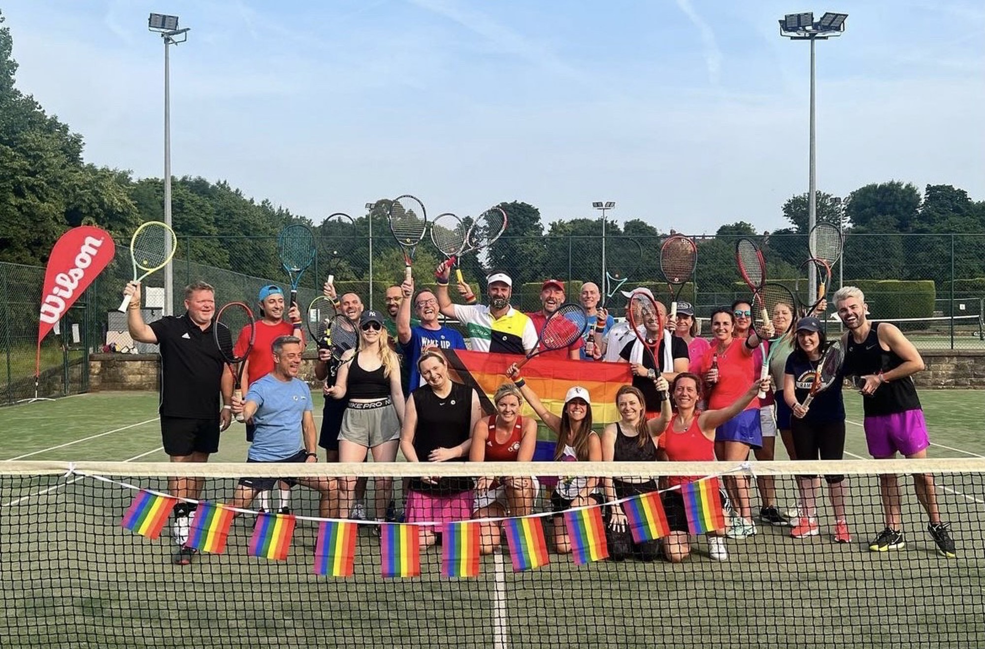 A large group of people standing on a tennis court, posing with tennis rackets dressed in brightly coloured clothing, behind a net draped with rainbow flags