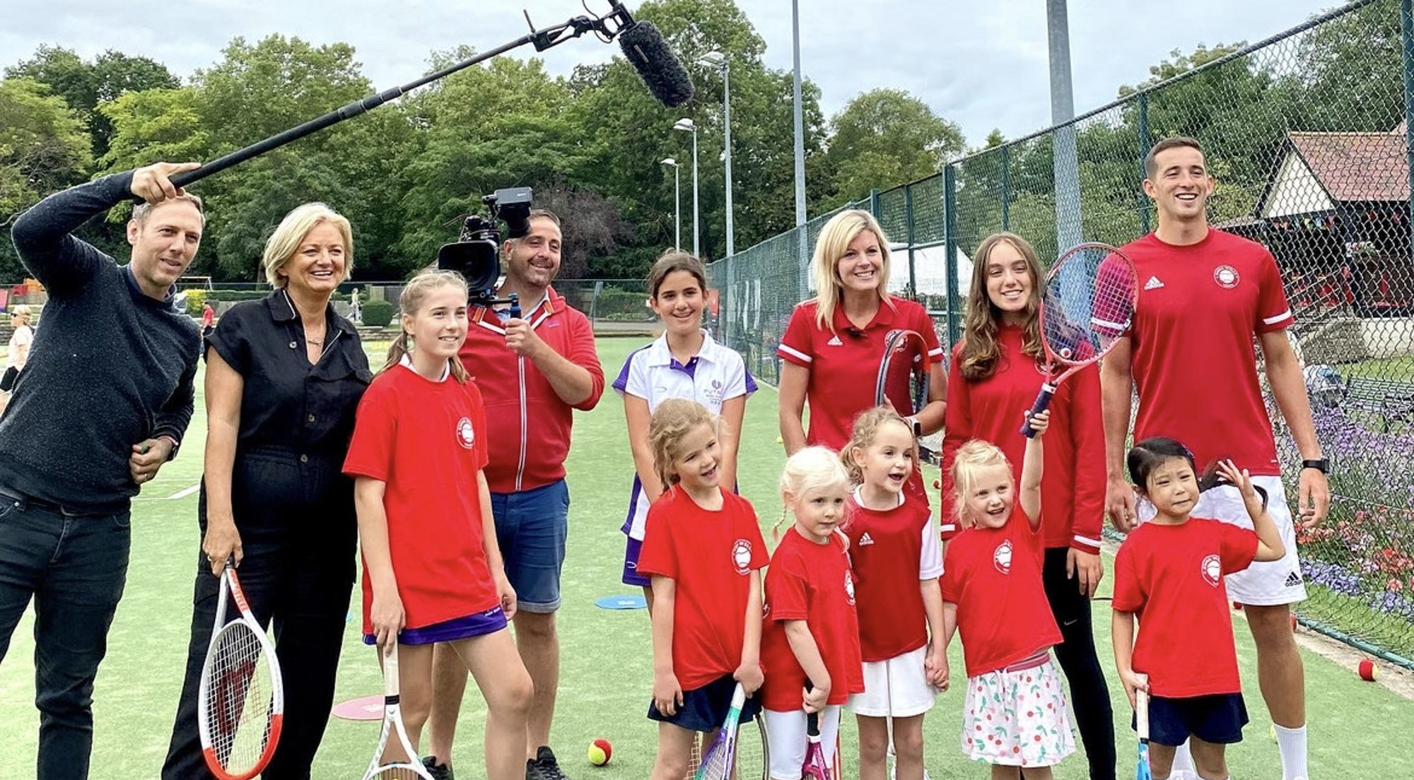 A group of children and adults posing with tennis rackets, with a man holding a large microphone and a man holding a tv camera stood next to them