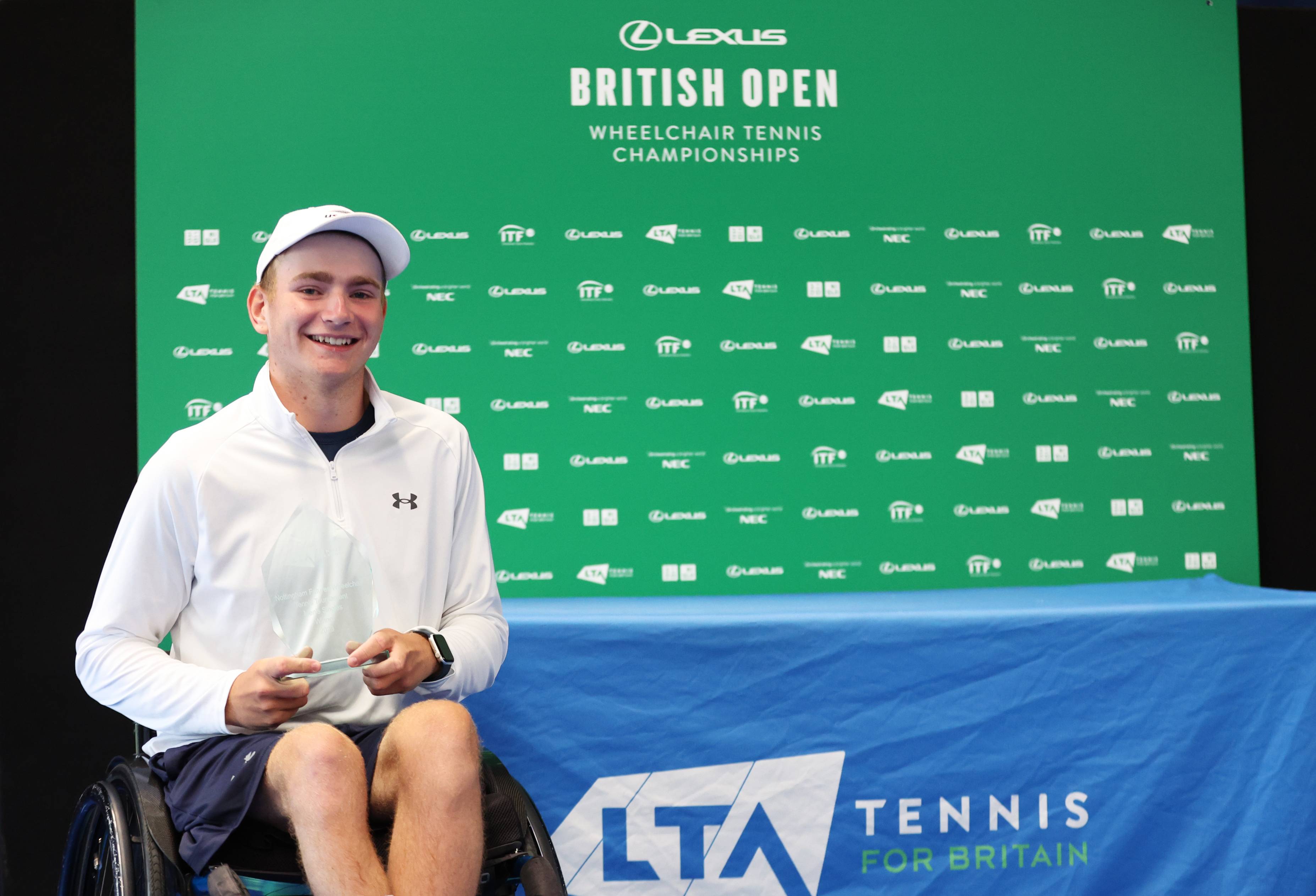 British wheelchair tennis player Andrew Penney smiling while sat in his wheelchair in front of a 'Lexus British Open Wheelchair Tennis Championships' branding board
