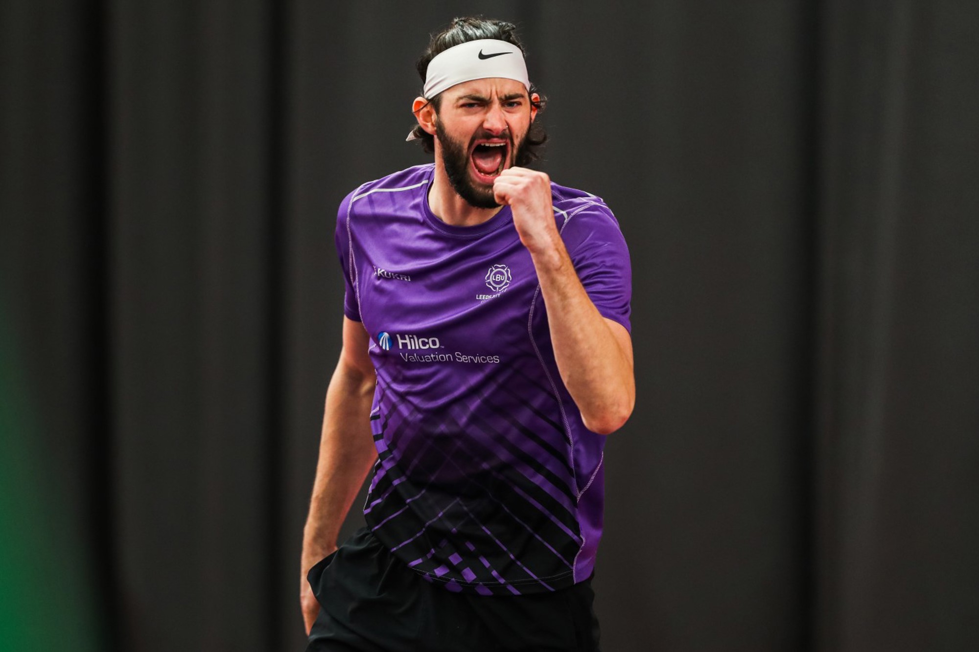 A man dressed in a purple shirt wearing a white headband clenches his fist in celebrating