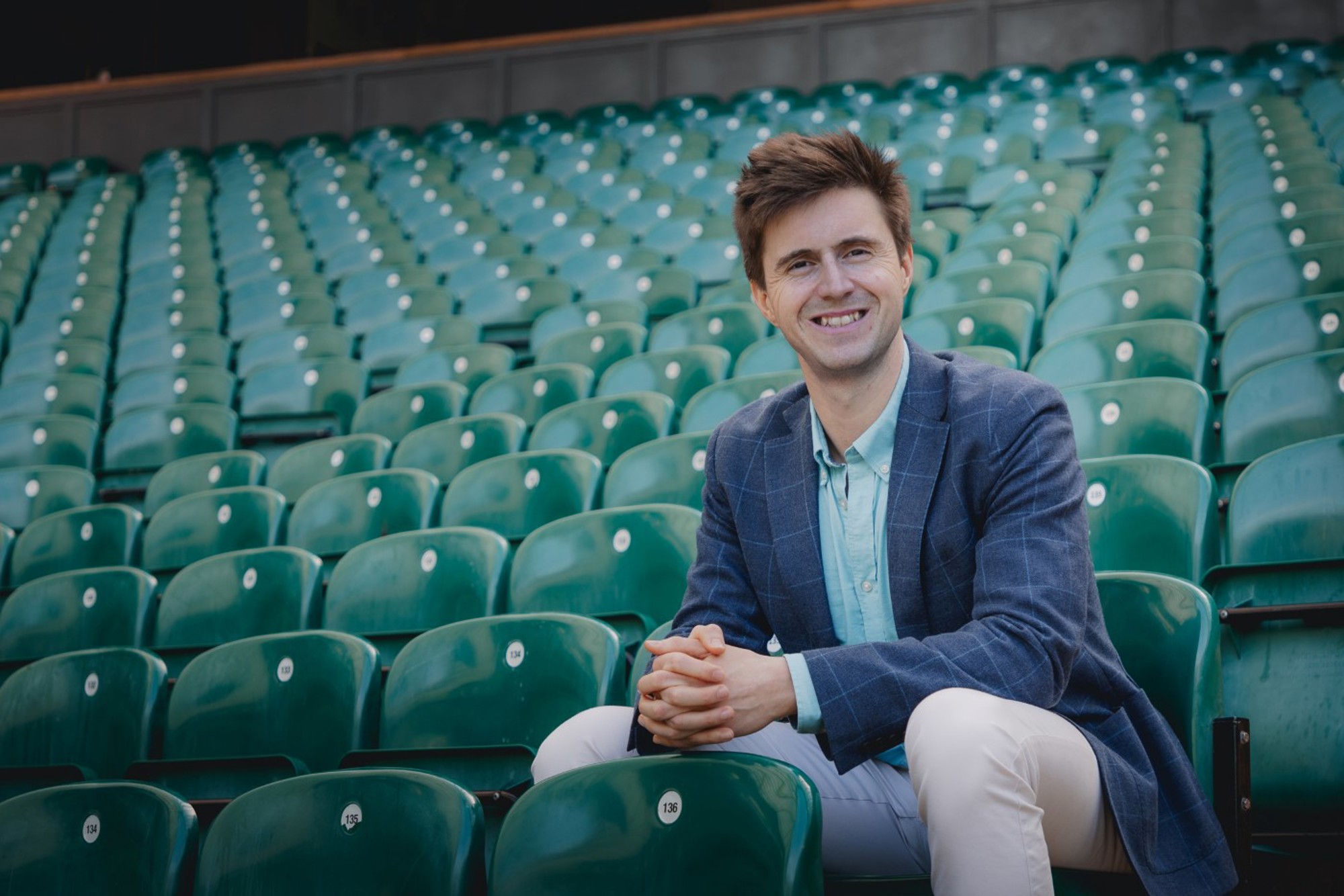 A man sat in green seating, clasping his hands together, wearing a blue checked blazer jacket
