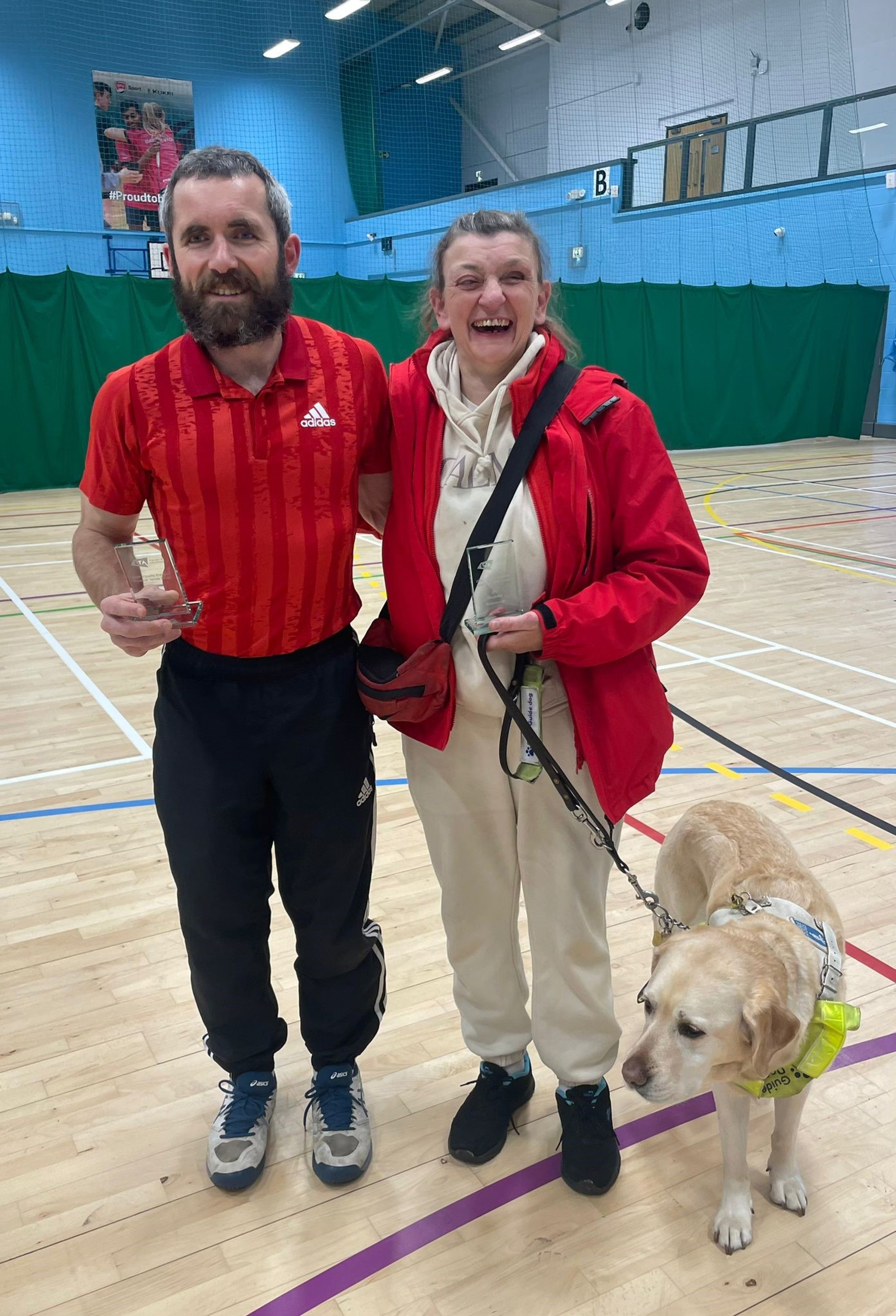 B1 doubles runners-up Roy Turnham (L) and Tracy Compton (R)  pictured with Guide Dog Sandy during the winner's ceremony at the 2024 Visually Impaired Tennis National Finals at Nottingham Trent University. 