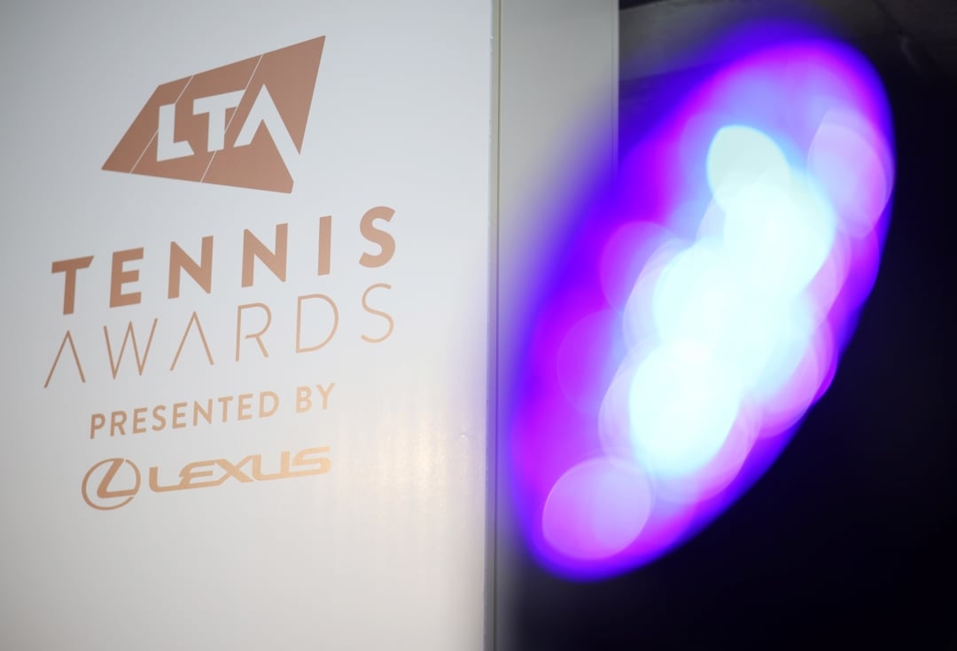 The text, 'LTA Tennis Awards presented by Lexus', in gold font on a white background, with a purple light out of focus to the right hand side