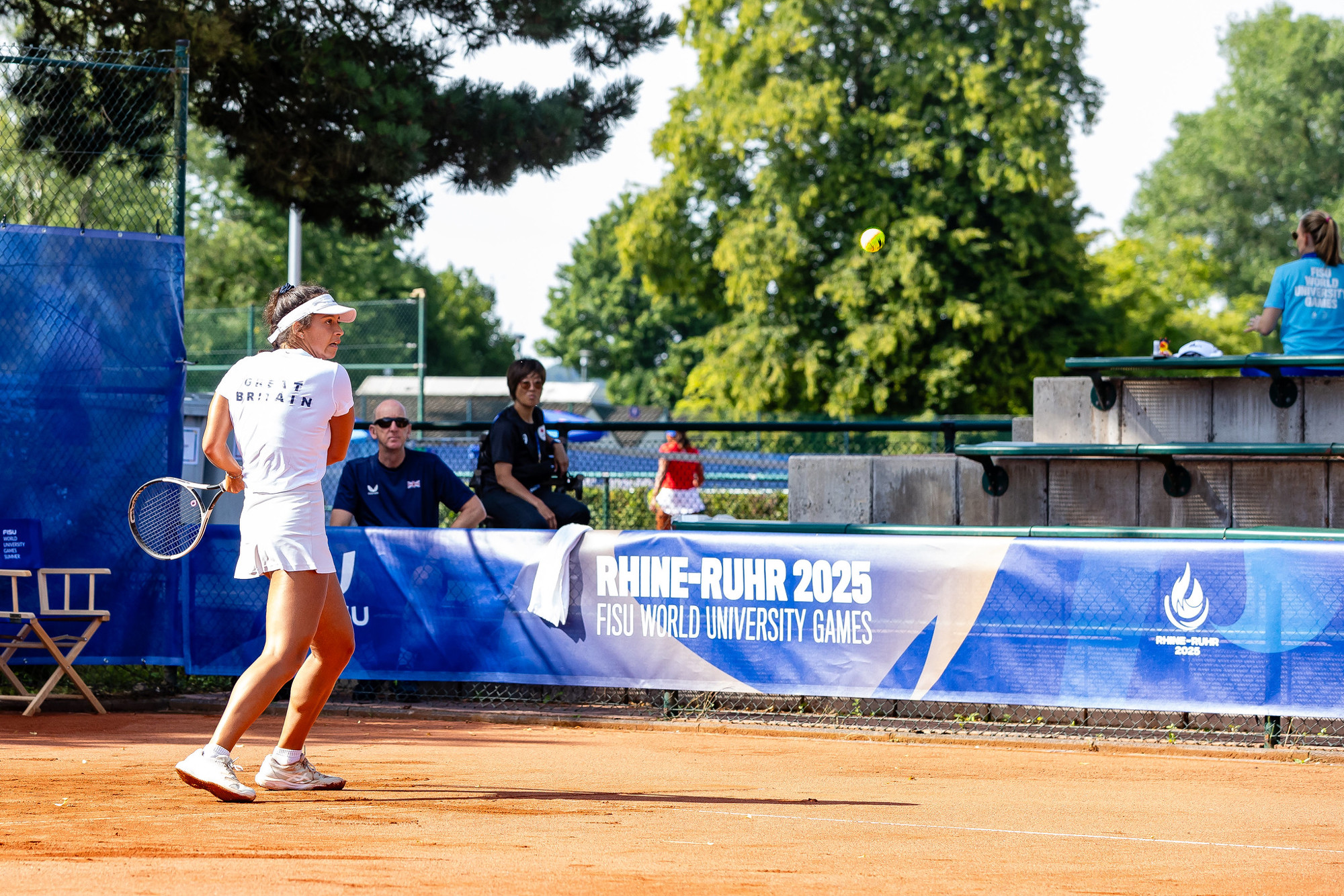 A woman wearing an all-white outfit consiting of a cap, shirt, skirt and shoes, stands on a clay tennis court preparing to hit a shot. A banner with the words 'Rhine-Ruhr 2025 FISU World University Games' is attached to a fence behind the player, while two people watch on from behind the fence, with trees visible further in the background. 