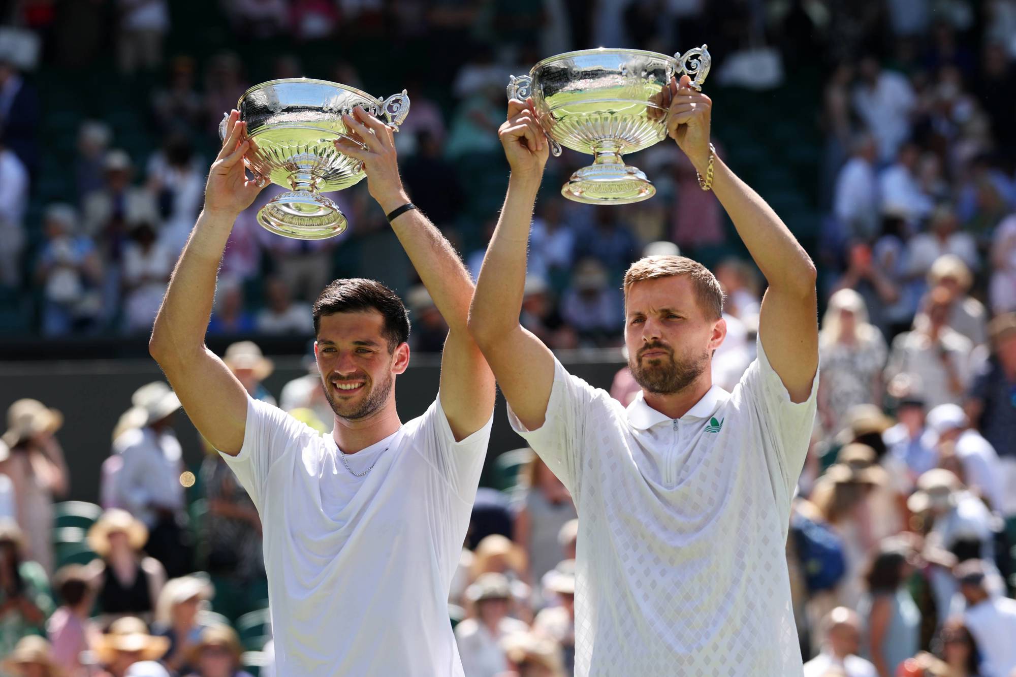 Julian Cash and Lloyd Glasspool smiling while holding up the Wimbledon men's doubles trophies above their head in front of the crowd