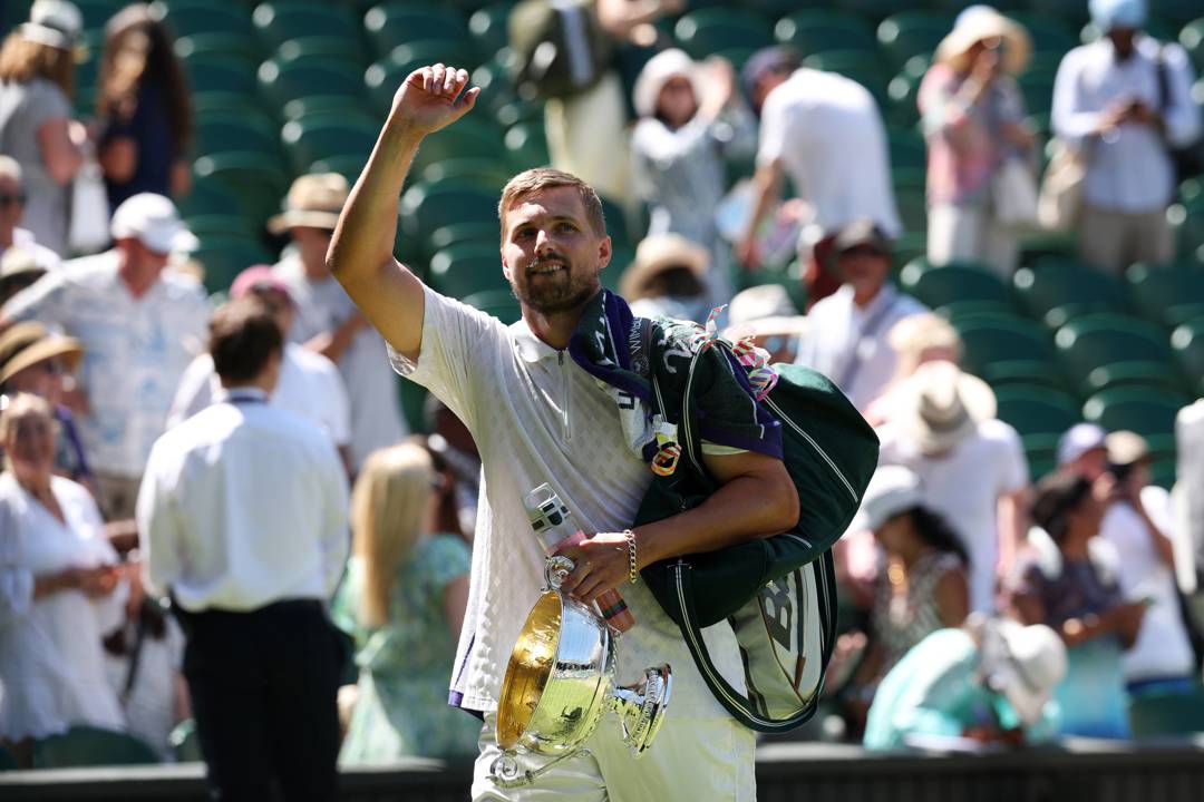 Lloyd Glasspool raising his hand to wave to the Wimbledon crowd, towel over his shoulder, holding onto his men's doubles trophy