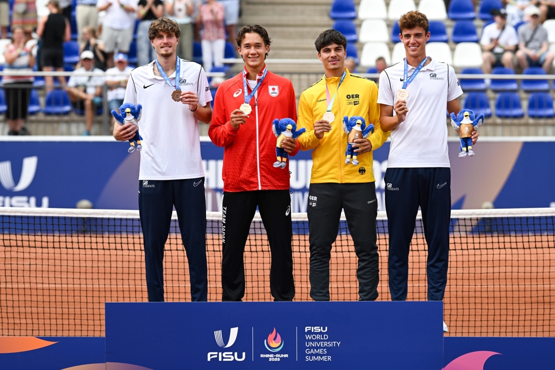 Four men wearing, from left to right, white, red, yellow and white sports tops, all showing of medals hung around their necks. A tennis net is directly behind them, and crowds are visible in stands in the background