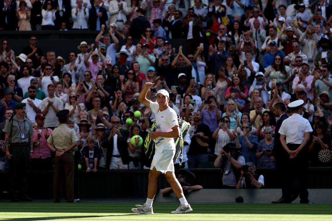 Oliver Tarvet, dressed in white, holding a thumb up to the crowd after his second round match against Carlos Alcaraz at Wimbledon 