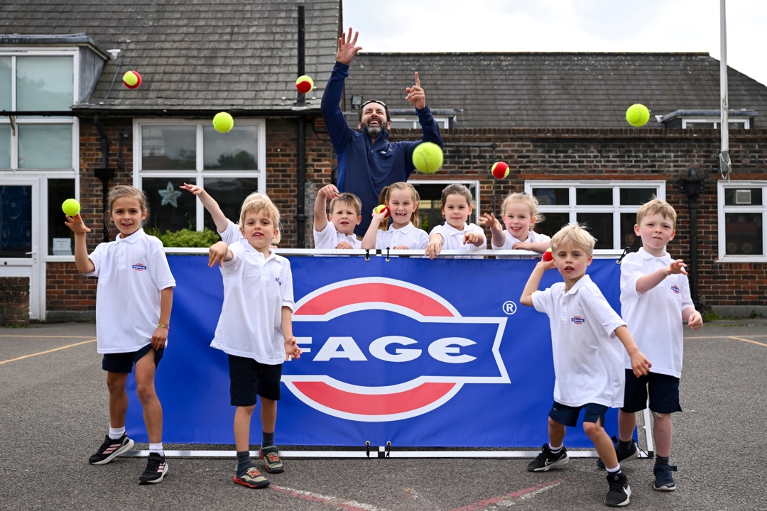 A group of children, stood in front and behind of a  blue banner with the word 'FAGE' on it, throw tennis balls towards the camera. There is an adult man stood with the group as well.