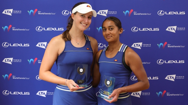 Two women wearing blue tennis dresses and holding glass trophies, stood in front of a blue background with sponsor logos from the LTA, ITF World Tennis Tour, and Lexus on display