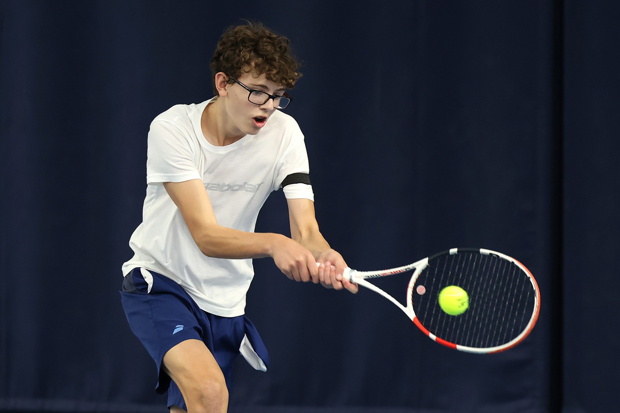 A boy wearing a white t-shirt, blue shorts and glasses hits a tennis balls. His mouth is slightly open with exertion