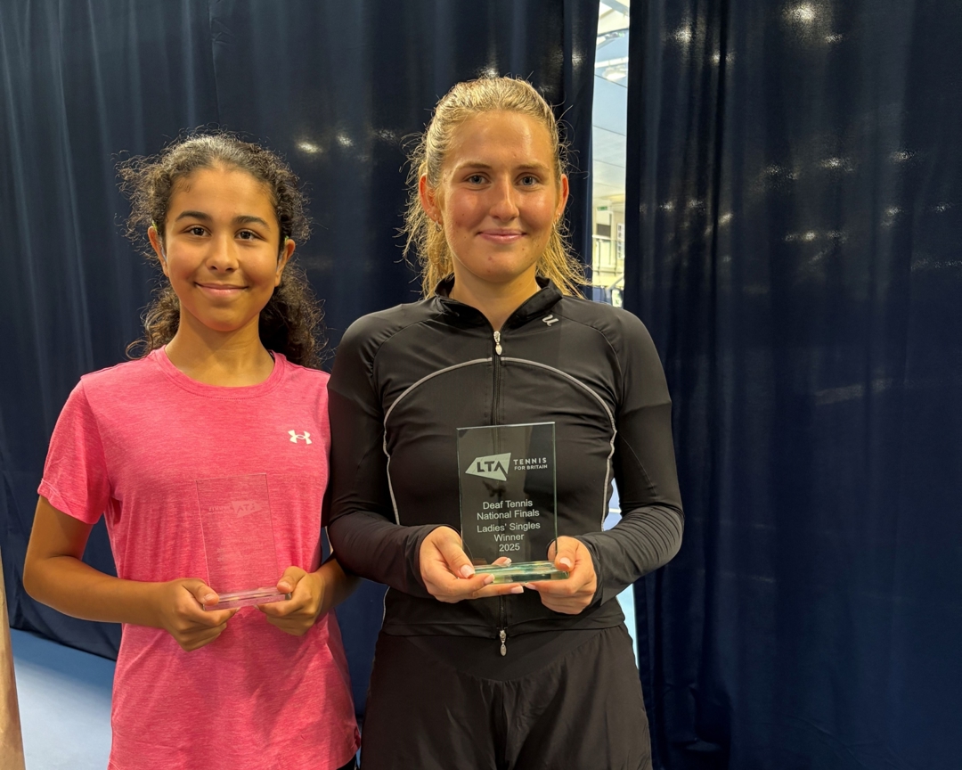 A girl in black short and a black t-shirt smailes at the camera, holding a glass trophy engraved with the words, 'Deaf Tennis National Finals Ladies' Singles Winner 2025'. Next to her stanbds a girl wearing a pink t-shirt, also holding a glass trophy and smiling