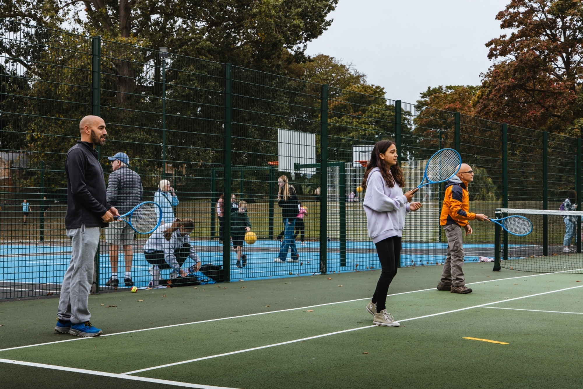 Three peopl two adult men and a young girl, each hold blue tennis racets in theire hands, while standing on a green tennis court. There is fencing behind them, spearting the tennis court from an area where adults and children are bouncing balls, and basketball hoops can be seen