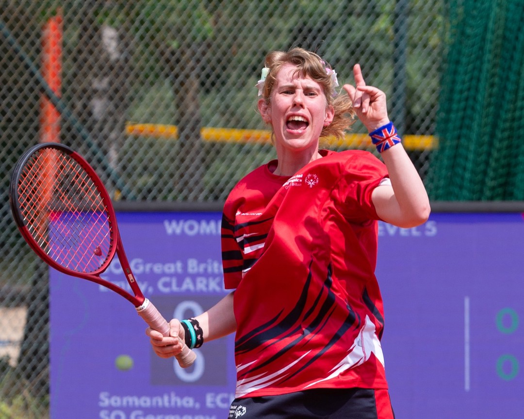 A girl in a red sports t-shirt celebrates, and holds a tennis racket in her right hand. On her left wrist, she is wearing a wristband with the British union flag on it.