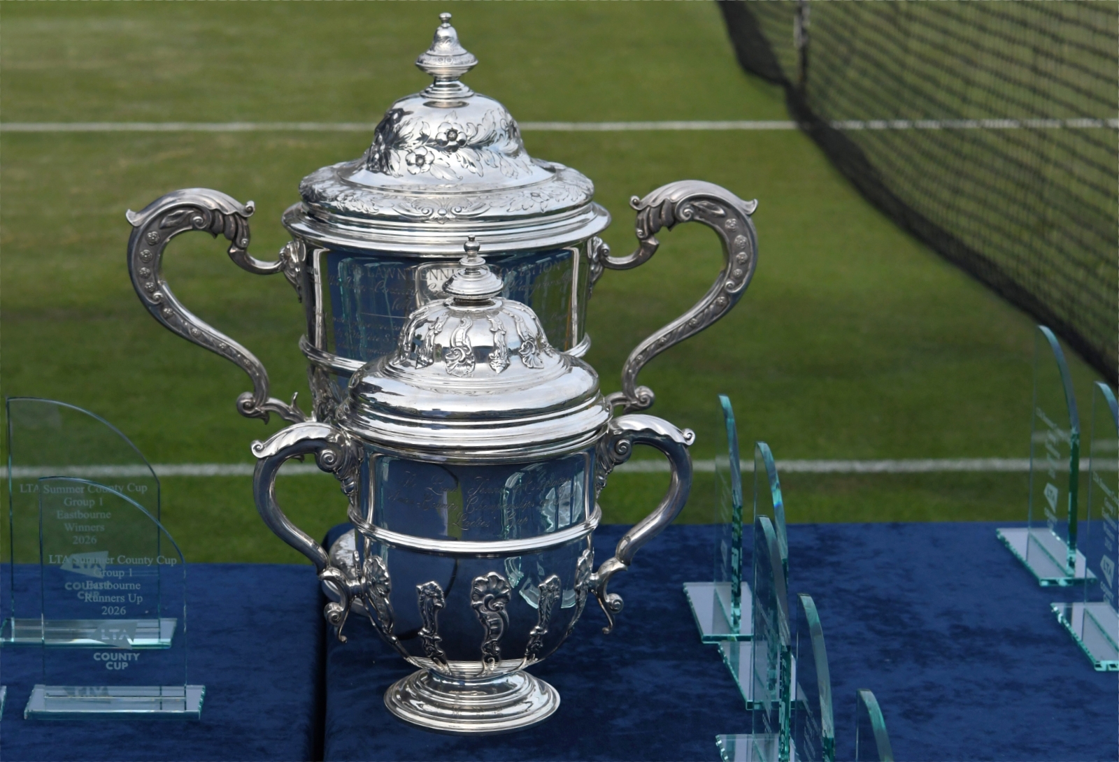Two large silver trophies sit on a table covered by a blue tablecloth, surrounded by smaller glass trophies. Behind the table, part of a grass tennis court is visible