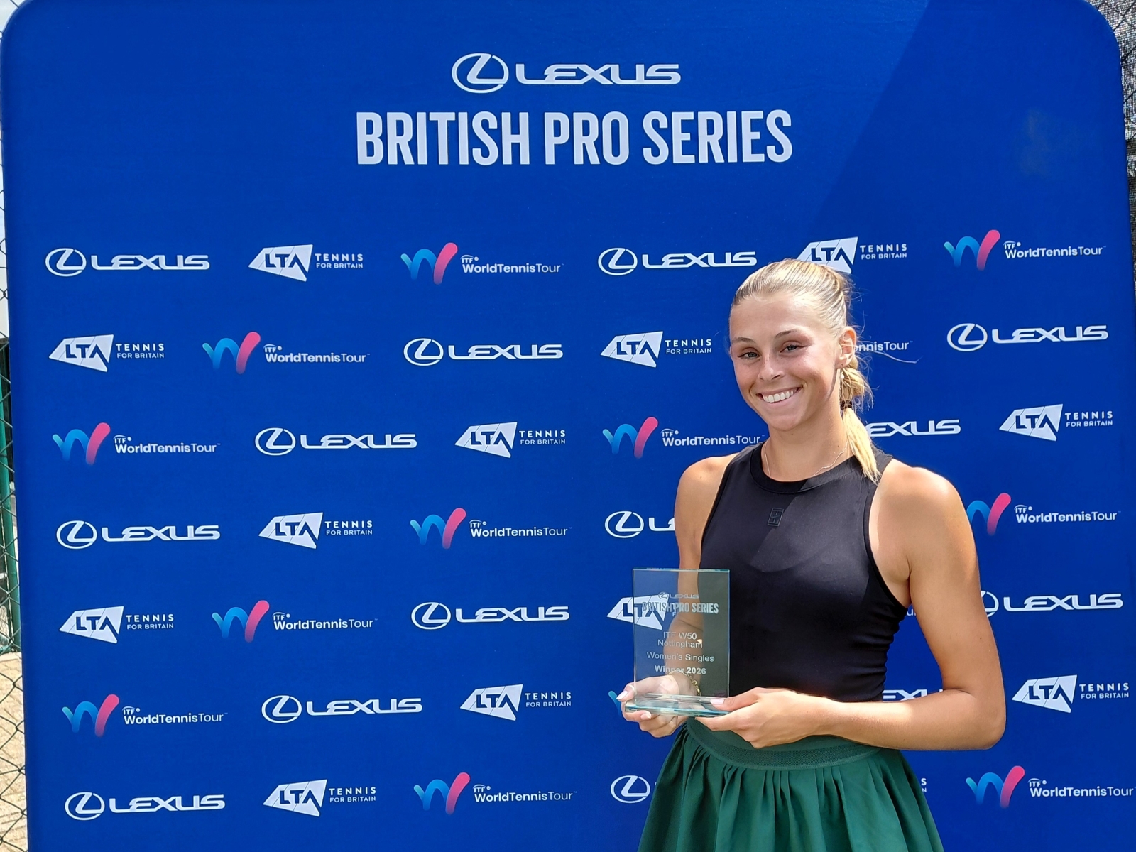 A woman wearing a black vest top and a green sports skirt holds a glass trophy, smiling. Behind her is a blue backdrop featuring the words, 'Lexus British Pro Series', along with a variety of sponsor logos