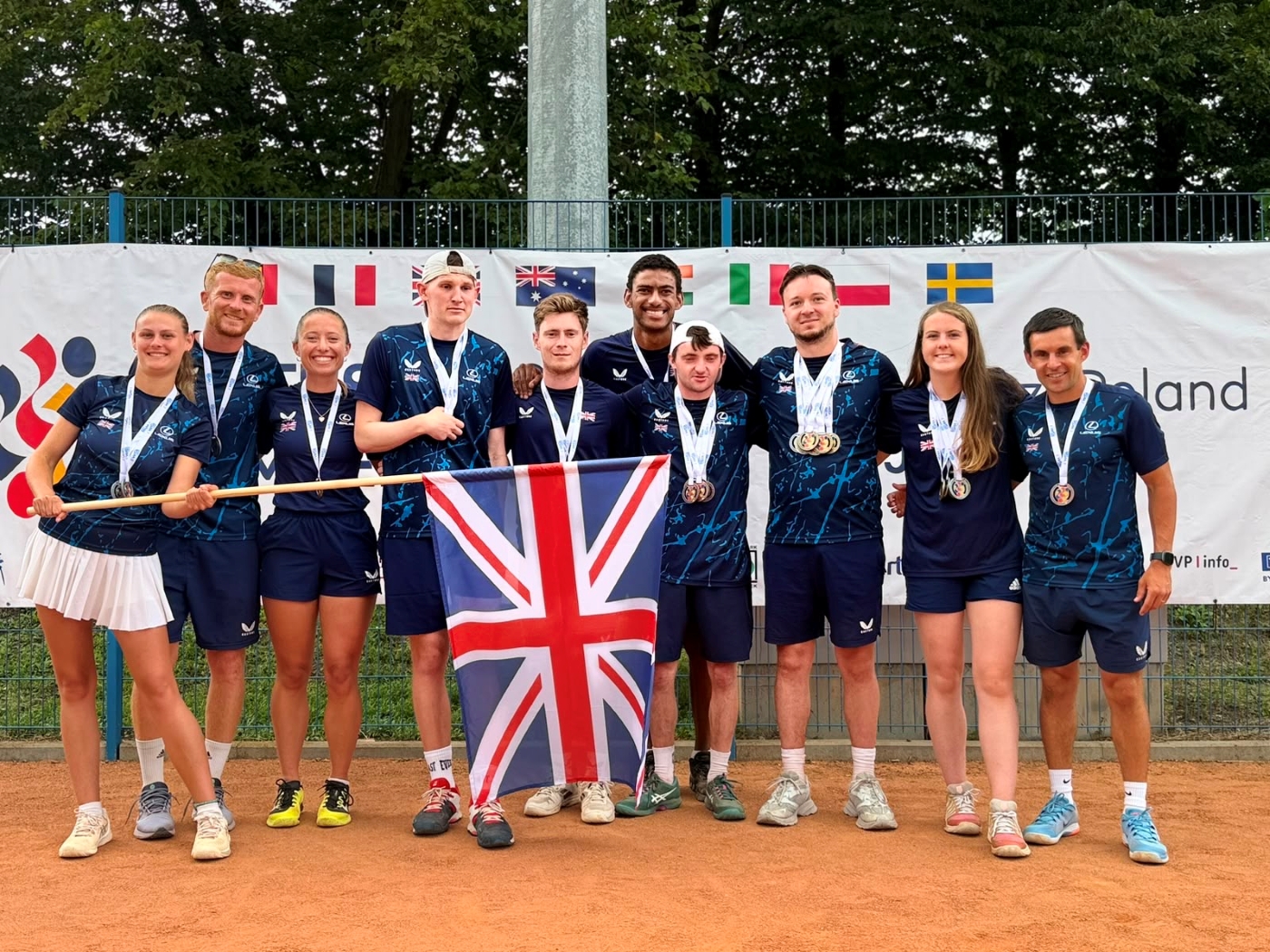 A group of people stand on a clay tennis court, with a British union flag being waved in front of the group
