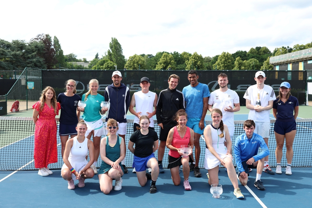 A group of men and women variously stand and kneel on a hard tennis court, with a woman wearing a pink vest top kneeling in the front row also holding a glass trophy