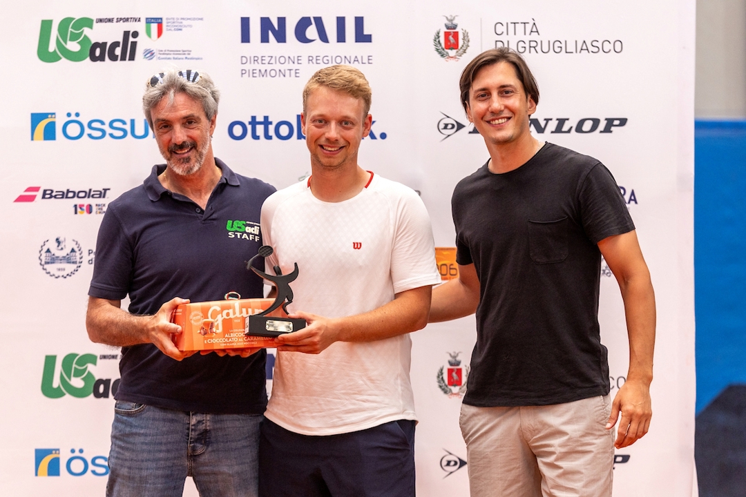 Three men stand side by side, with the man in the middle, wearing a white-t-shirt and black shorts, holding a black trophy