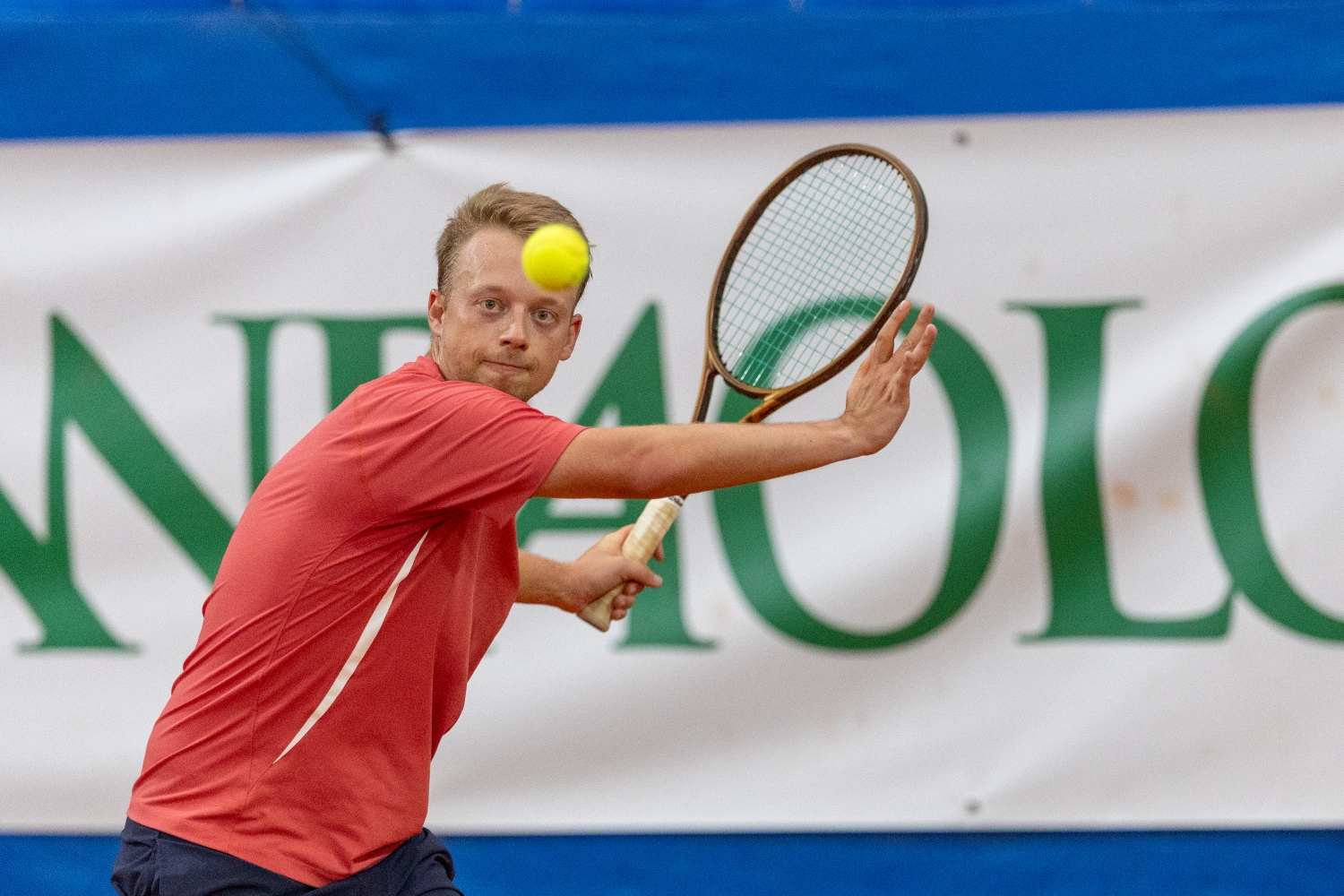 A man wearing a red t-shirt prepares to hit a tennis ball with a tennis racket. Behind time, a white banner with green letters on it is visible, with strips of blue present above and below the white banner