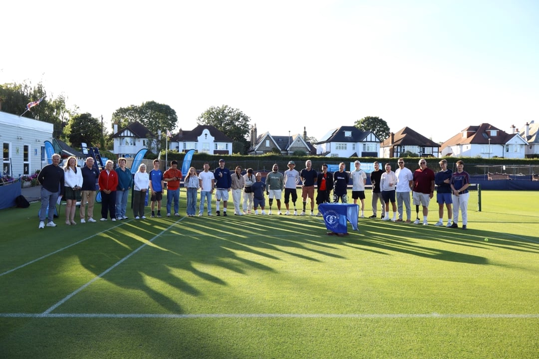 A group of people stand on a grass tennis court. Long shadows are being cast on the floor.