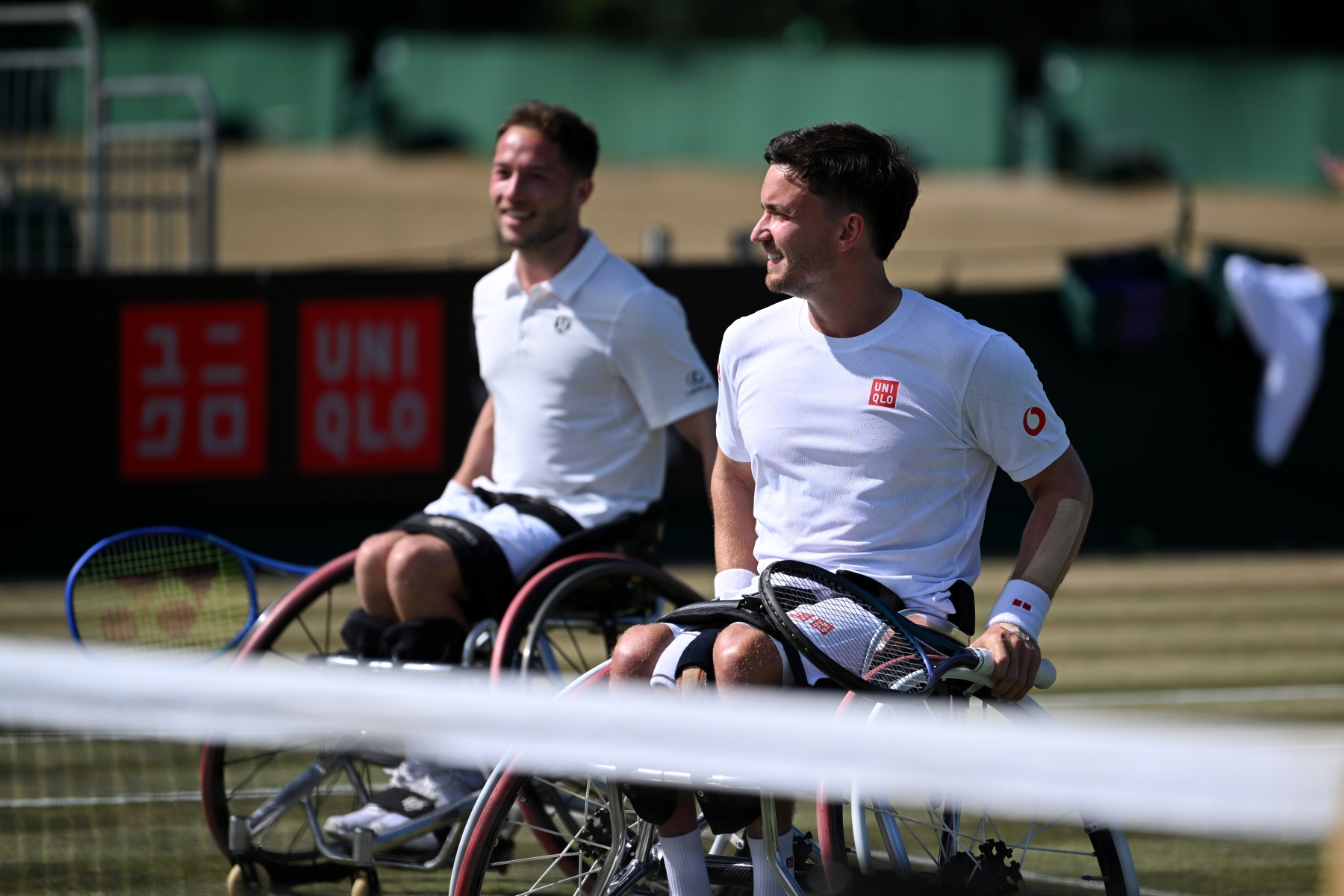 Two men in wheelchairs wearing white t-shirts and shorts and holding tennis rackets smile as they wheel themselves towards a tennis net