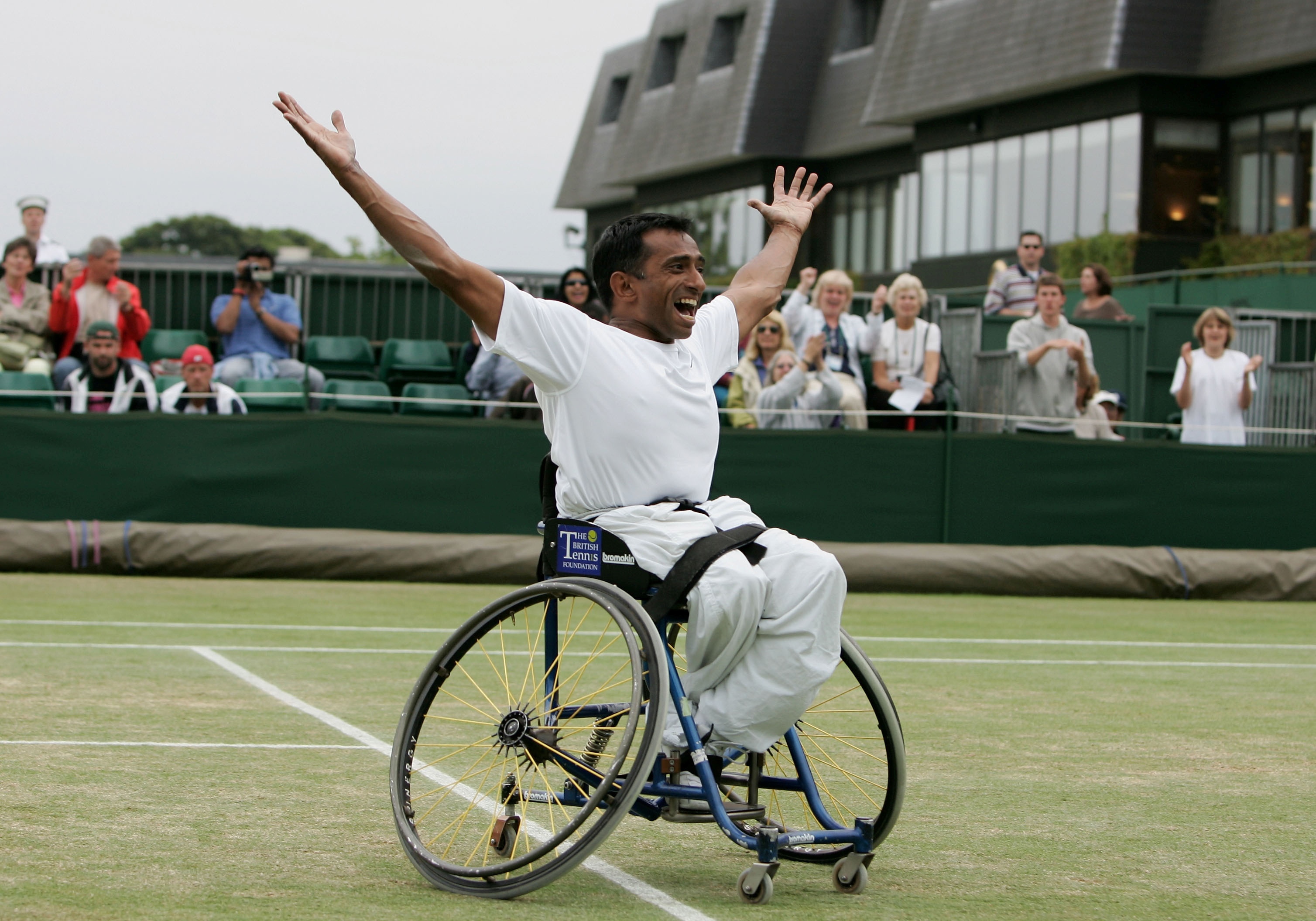 A man in a wheelchair wearing all-white clothing whilst on a grass tennis court throws his hands in the air in celebration