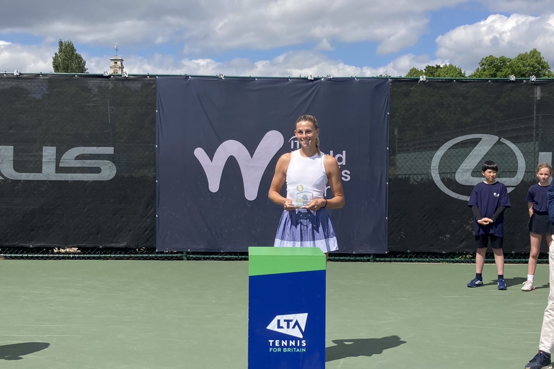 A woman wearing a white vest top and a lilac skirt holds a glass trophy, standing behind a blue pedestal with the words, 'LTA, Tennis for Britain' on it