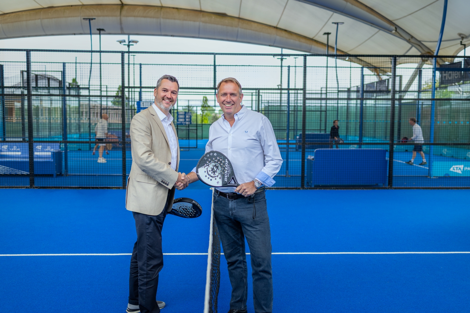Two men stand on a blue padel court, shaking each other's hand and each holding a padel bat in the other. People on another padel court are visible in the background.