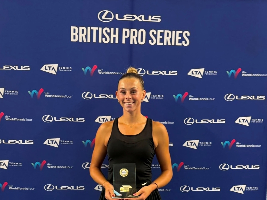 A woman wearing a black vest top satnds smiling and holding a glass trophy. Behind her is a blue backdrop featuring a variety of sponsor logos, with the words Lexus British Pro Series at the top