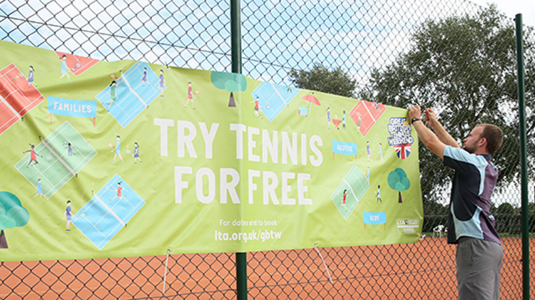 Male player attaching banner to courtside fence, that reads 'Try Tennis for Free'