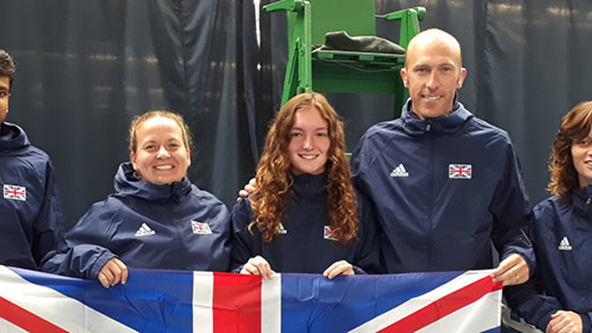 four tennis players and coach wearing GB blue jackets holding up  big GB flag