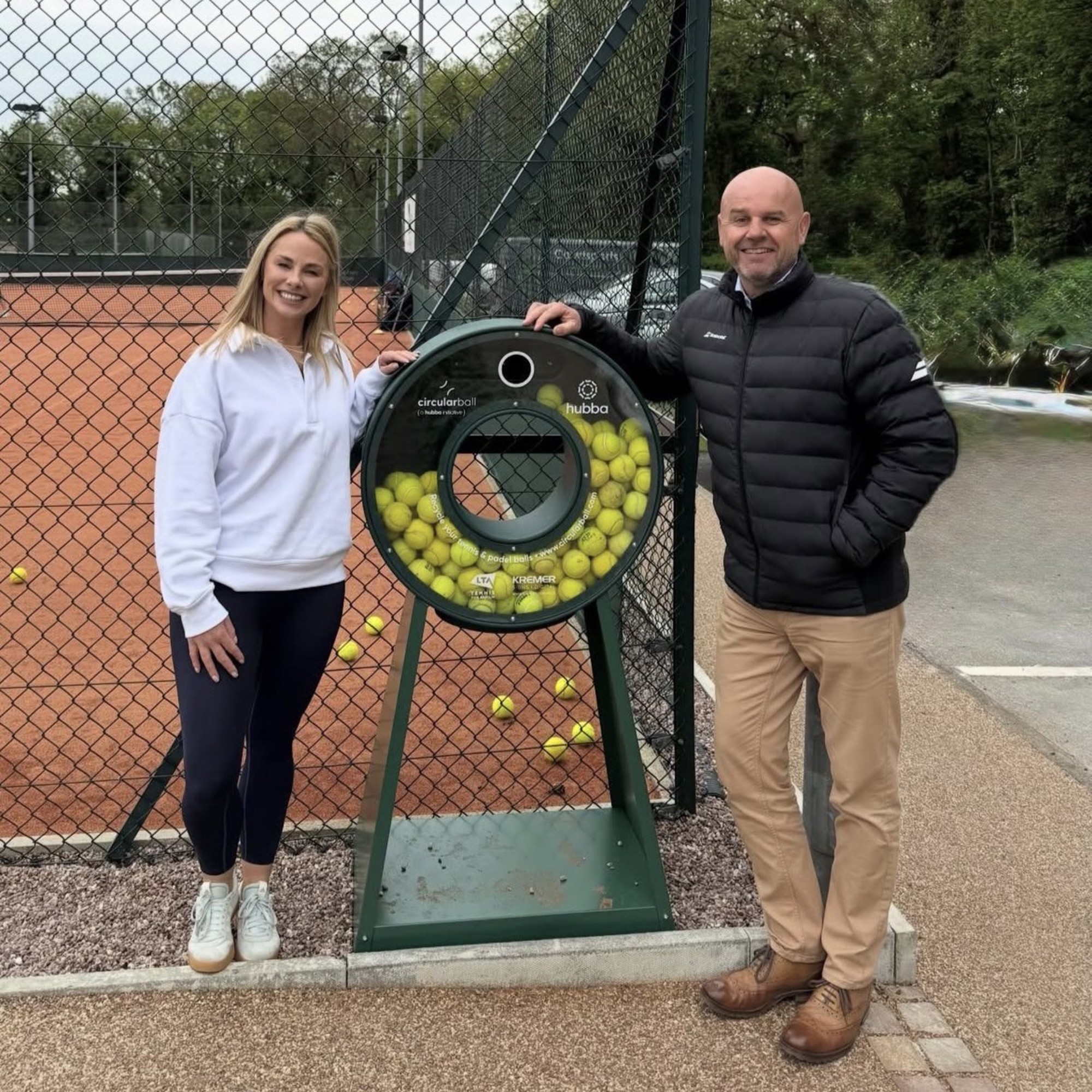 A man and woman posing next to a tennis ball recycling point next to a tennis court.
